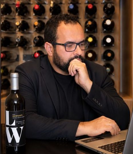 Lussandro Ilha em uma adega, diante do notebook, ao lado de uma garrafa de vinho